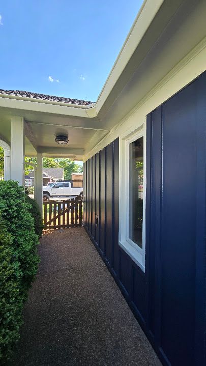 Narrow outdoor side passage beside a blue house, leading to a wooden gate and bright backyard.