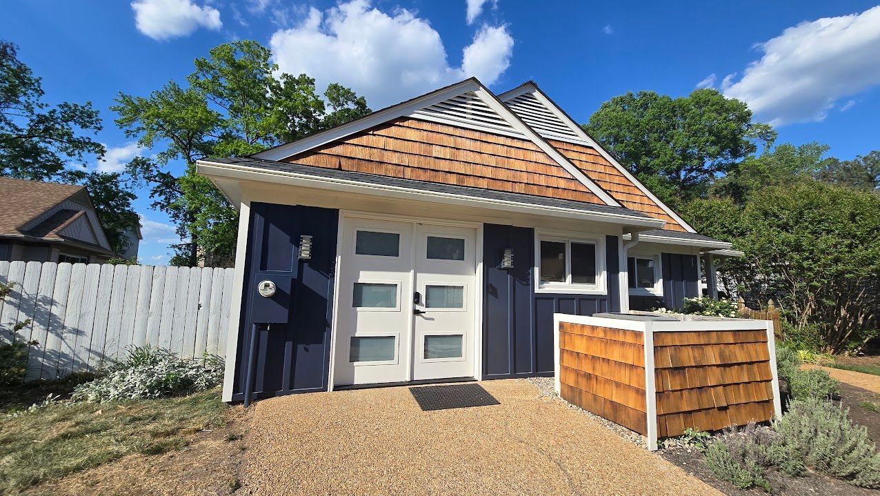 Small white cottage with tan shingled roof, front porch, and gravel yard under a blue sky.