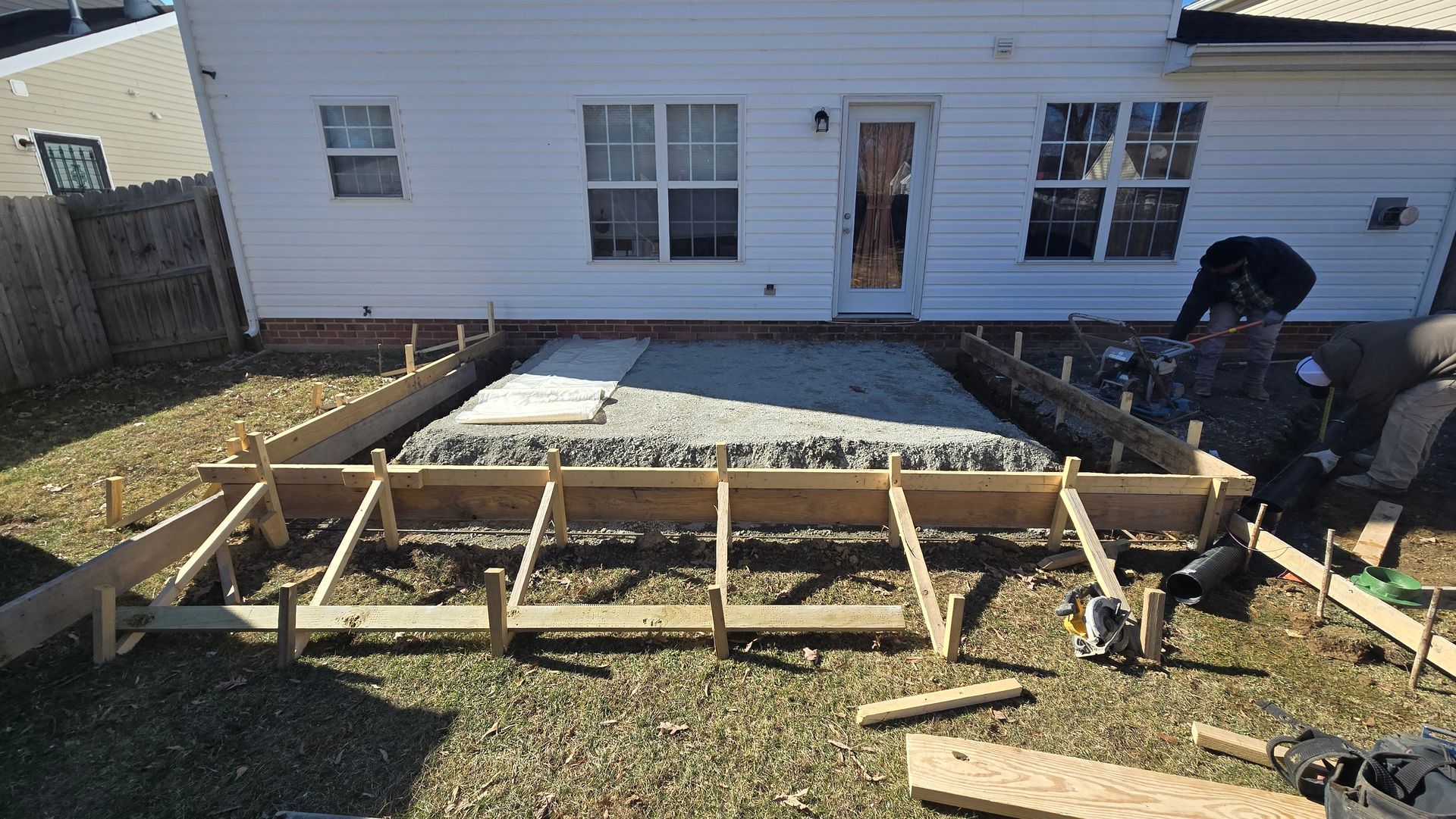 Backyard deck framing under construction beside a white house, with tools and lumber on the ground.