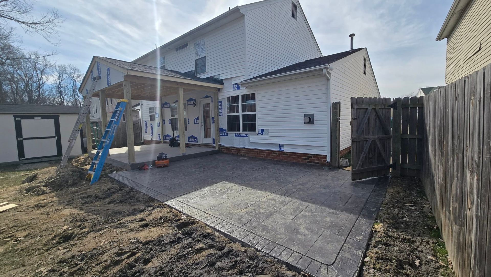 Backyard patio under construction beside a white house, with concrete slab, dirt yard, and wooden fence