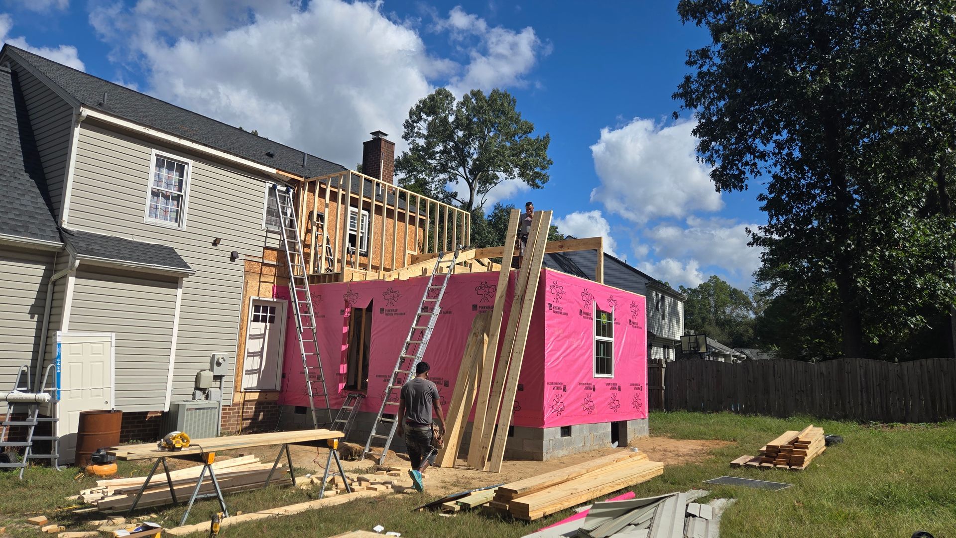 House under construction with pink siding, ladders, lumber, and scaffolding in a backyard under a sunny sky