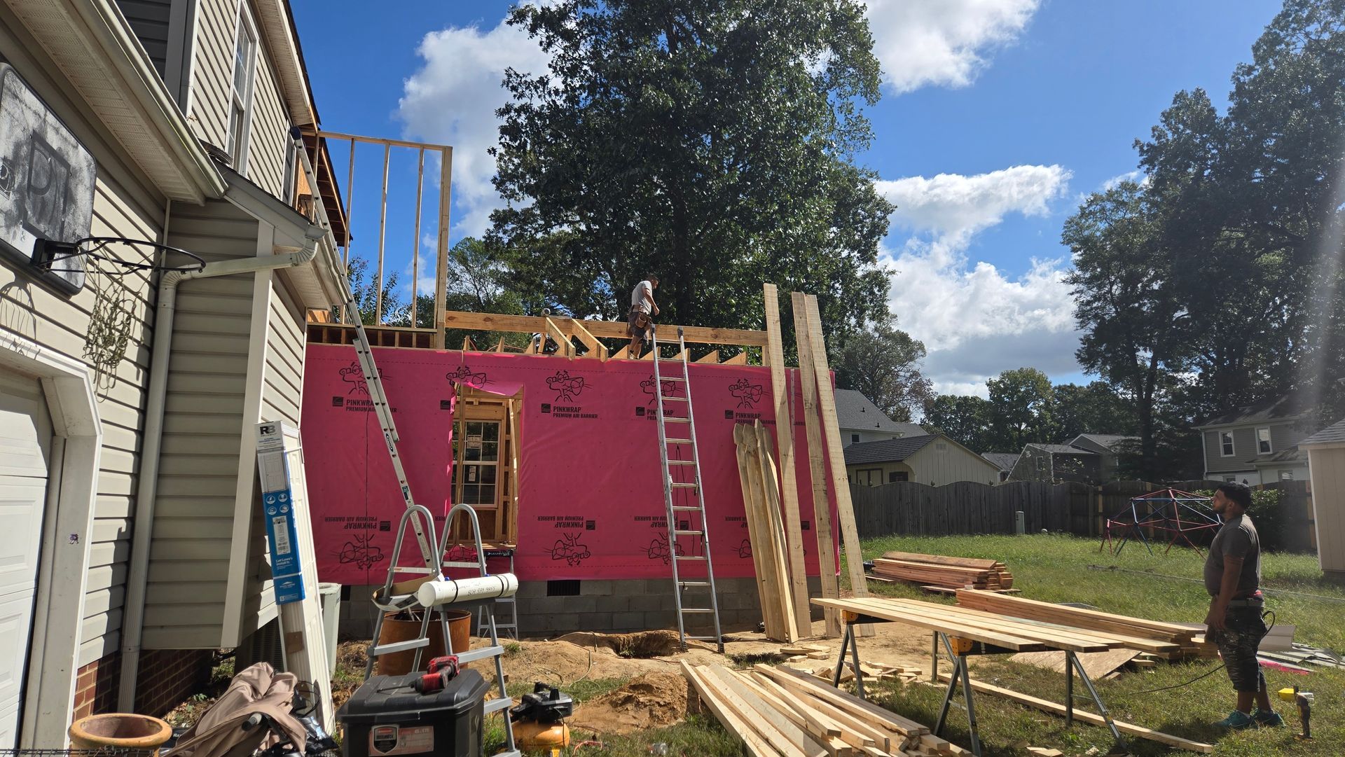 Workers framing a house addition with pink sheathing and lumber beside an existing home