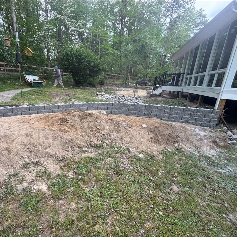 Backyard construction site with a retaining wall, dirt slope, and screened porch beside trees