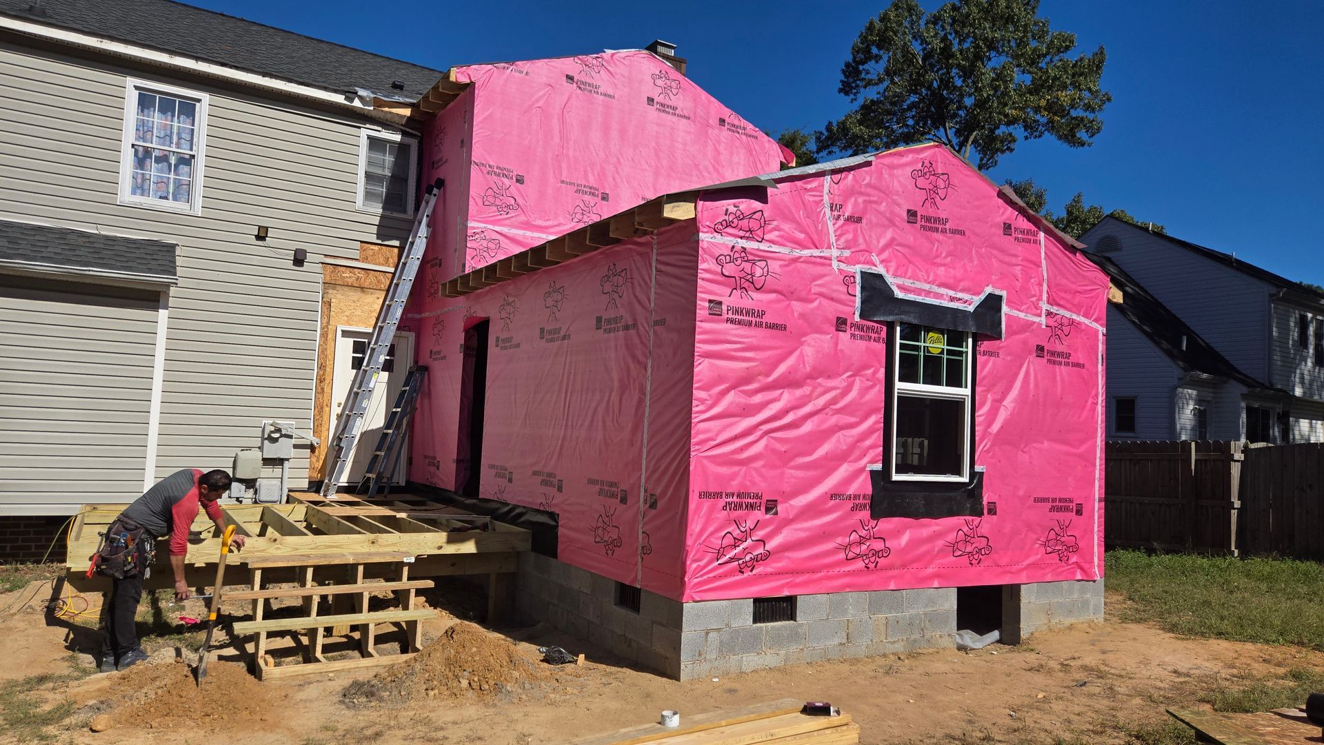 House under construction wrapped in bright pink house wrap, with a worker building a wooden deck nearby.