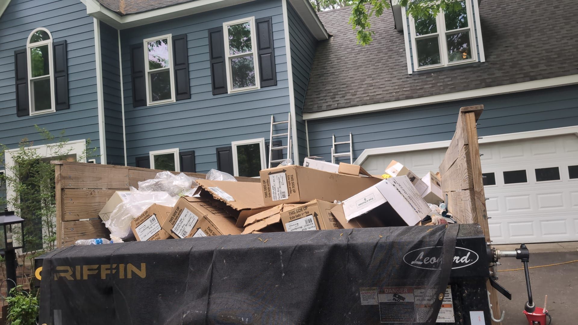 Black dumpster full of construction debris in front of a blue house and garage