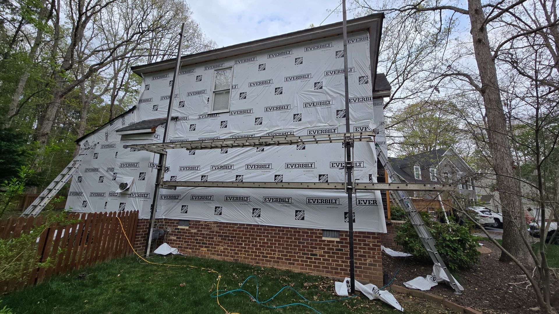 House exterior with unfinished gray siding and exposed brick foundation beside wooded yard