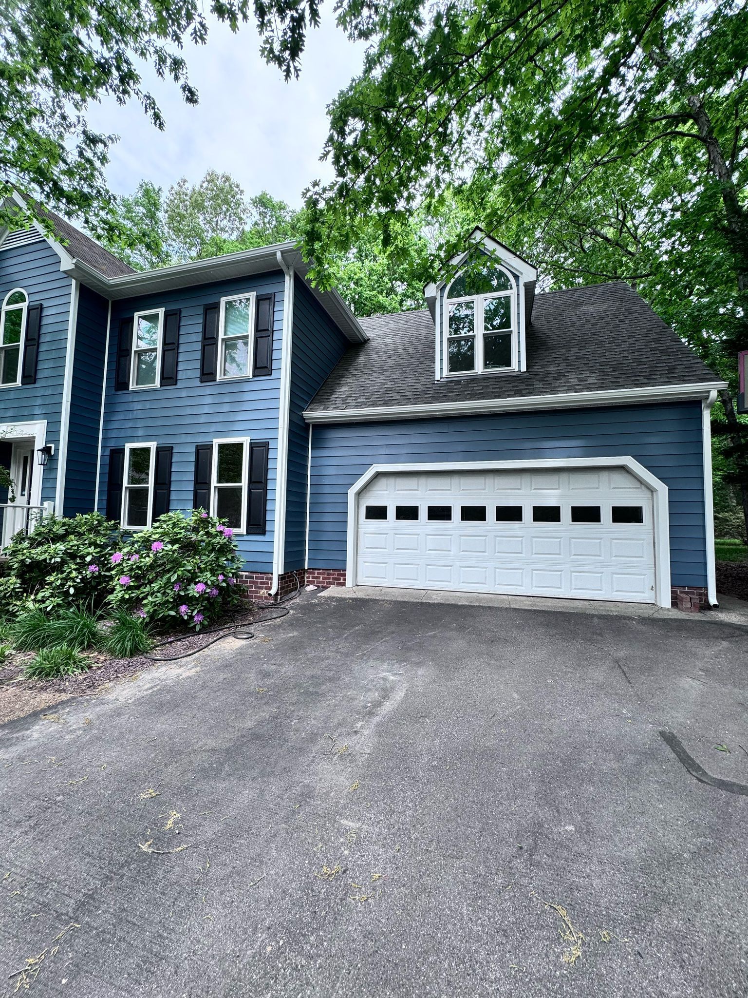 Blue two-story house with white garage and driveway, framed by trees and shrubs