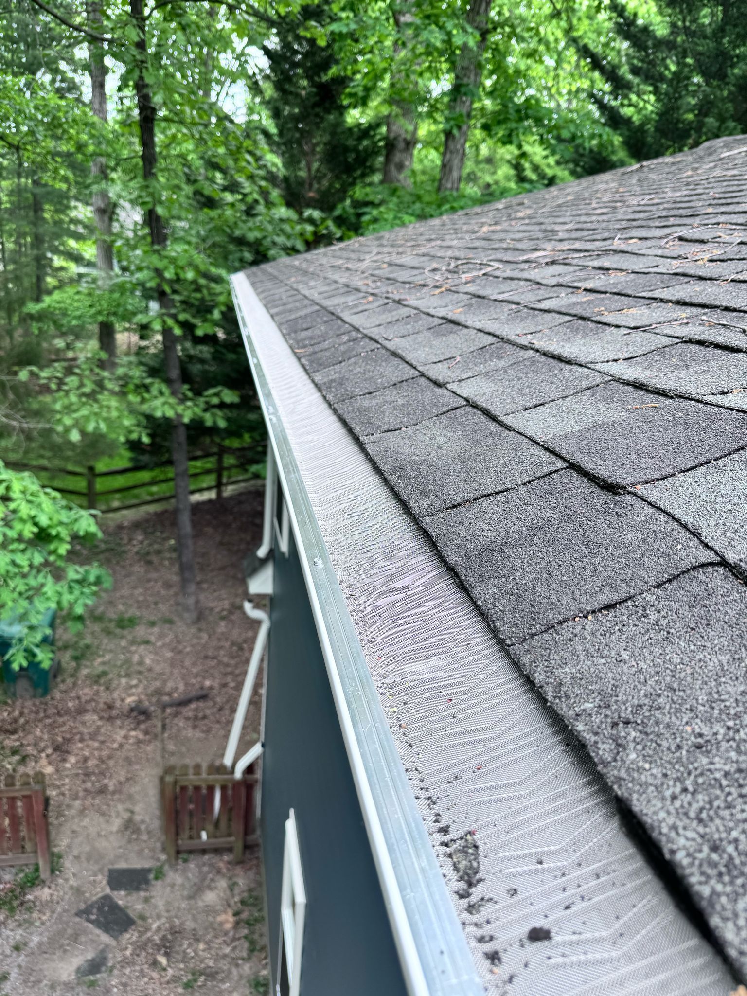 View of a gray shingled roof beside trees and a wooded yard from above