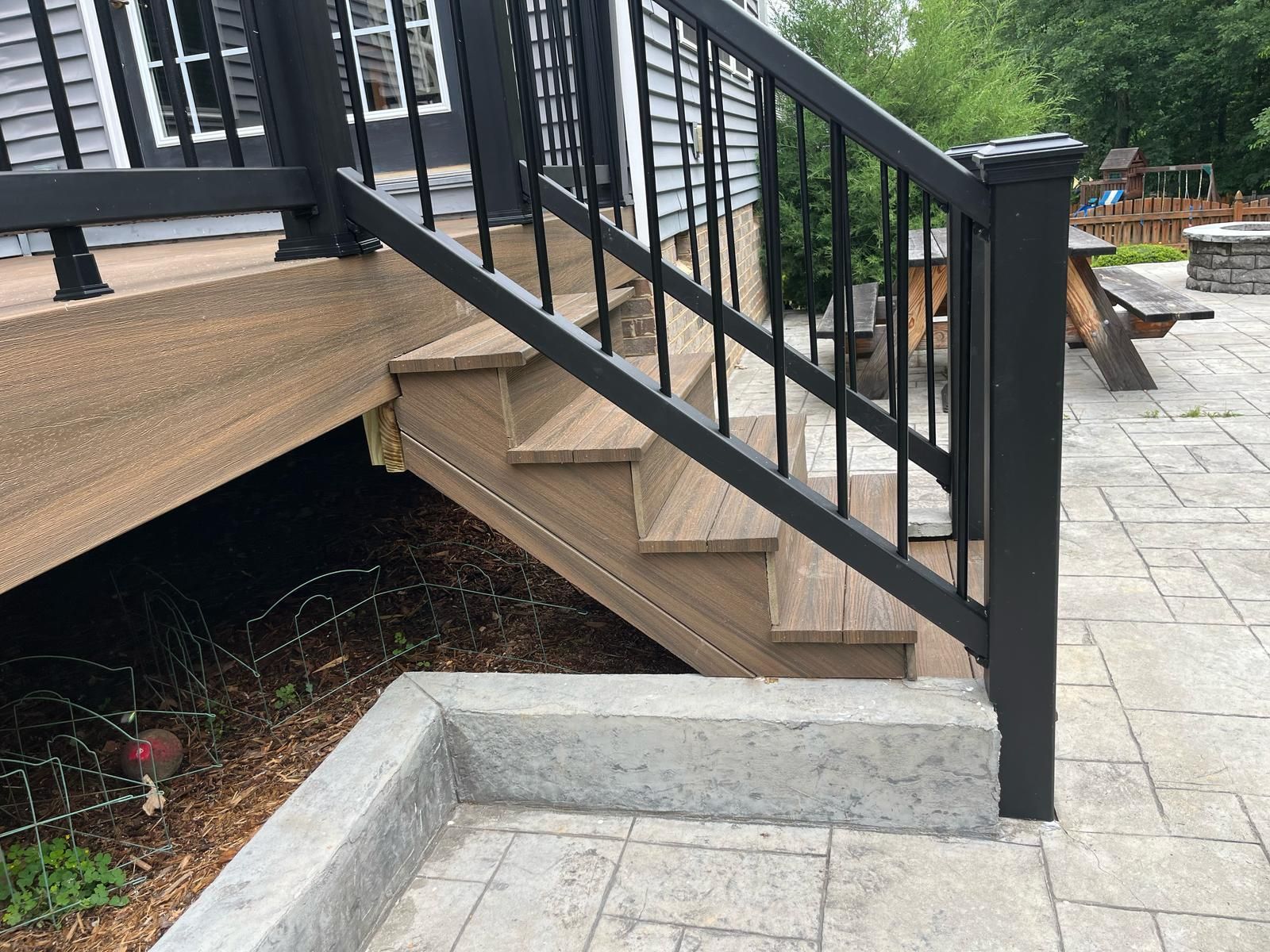 Concrete steps and black railing beside a deck, with a small ramp leading to a paved patio.