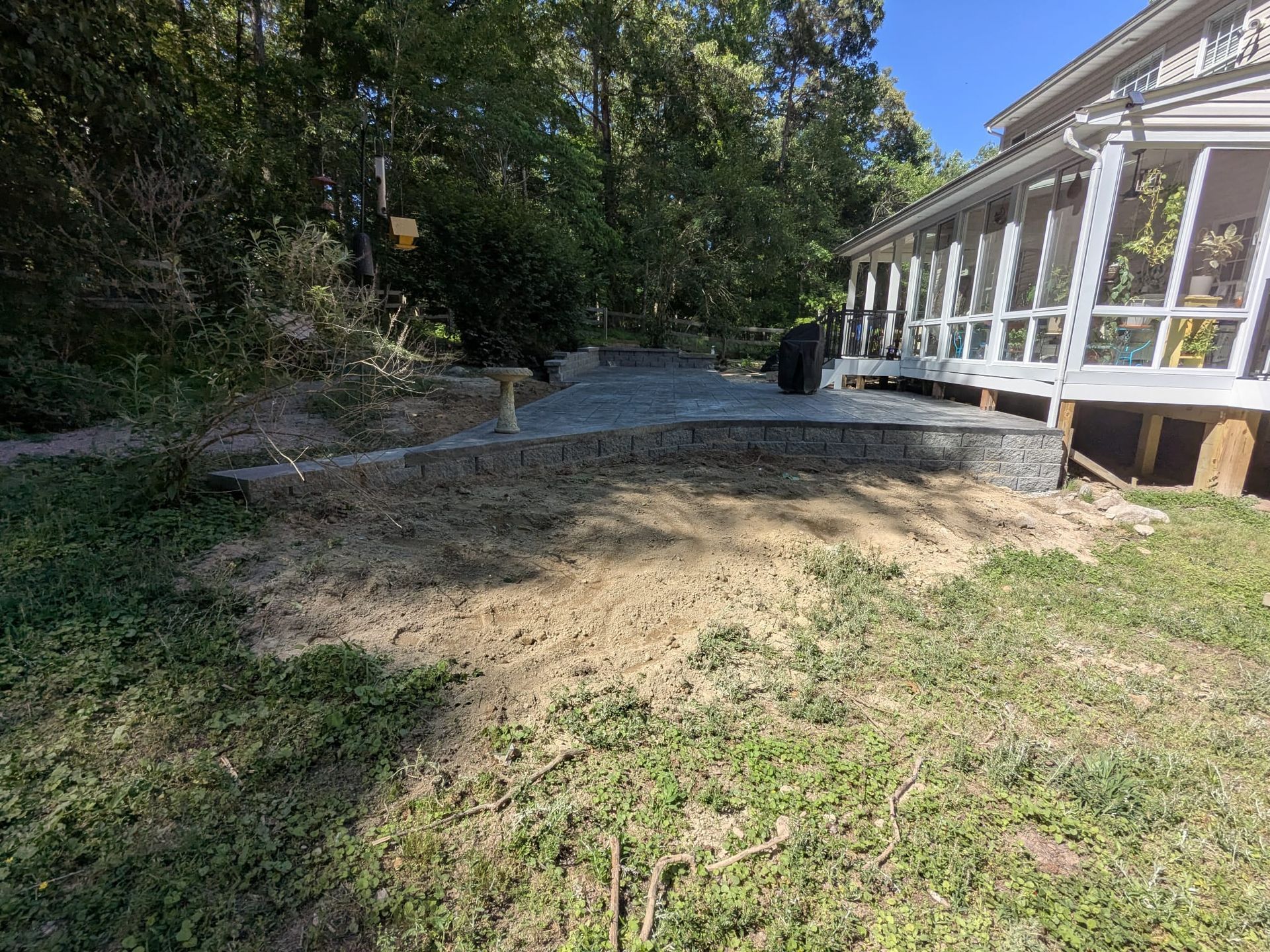 Dirt path beside a white porch, leading into a wooded yard under bright daylight.