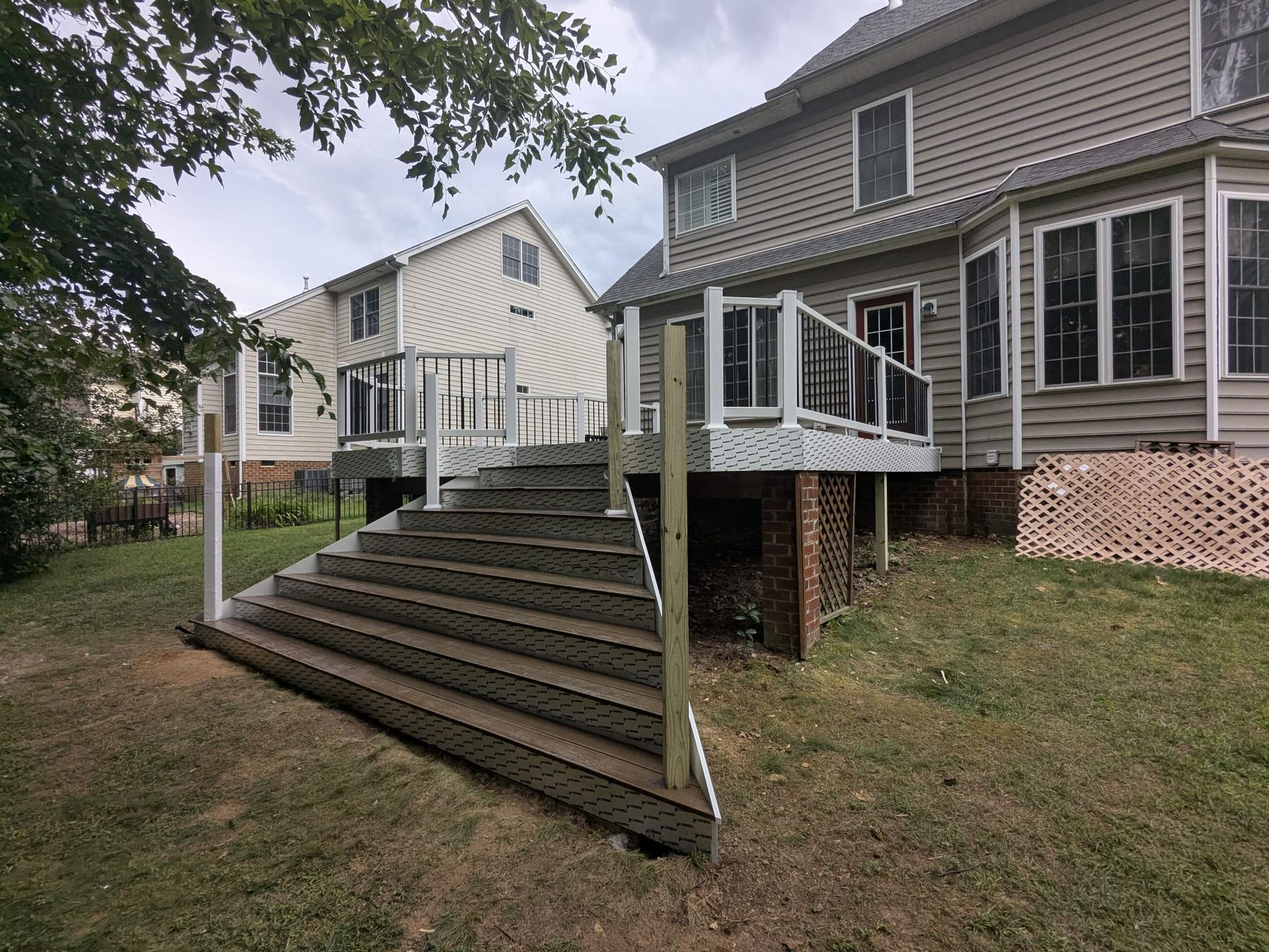 Backyard deck with wide wooden stairs beside a gray house and white outbuilding under trees