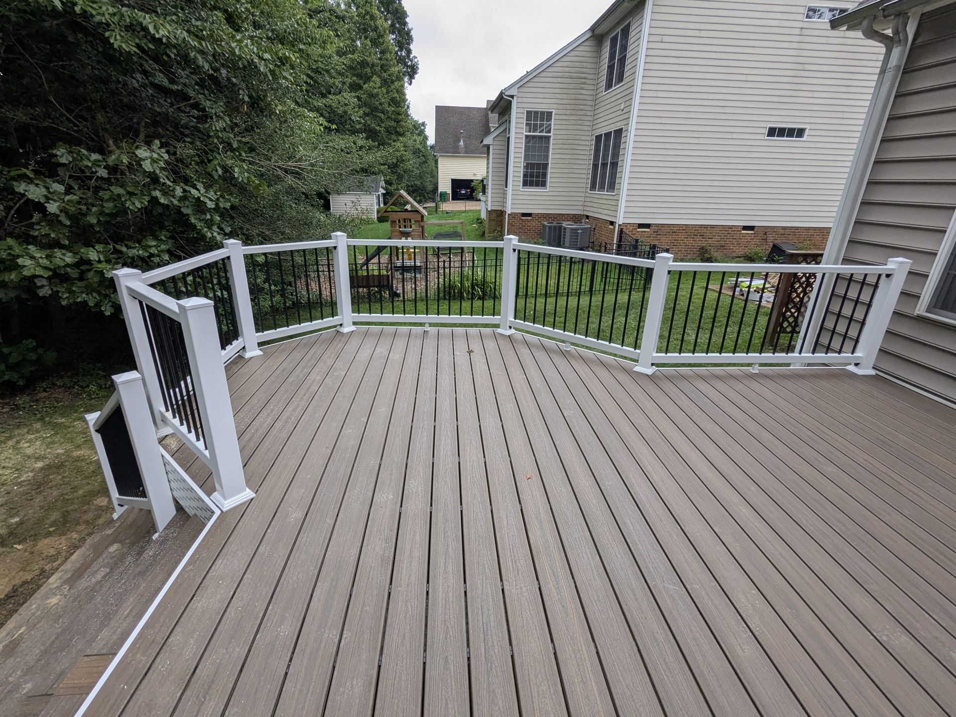 Backyard wooden deck with white railings and steps, beside a house and trees.