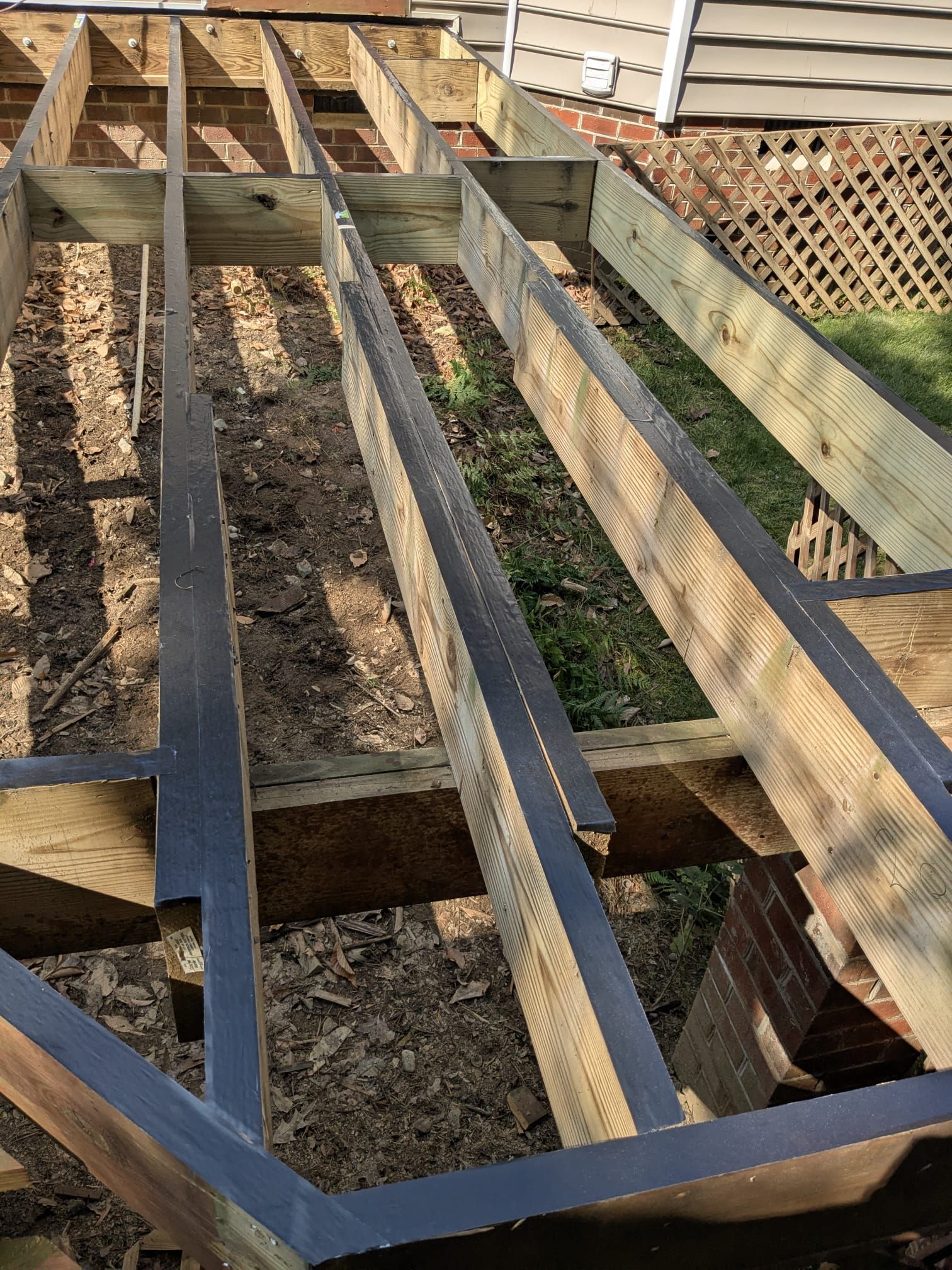 Wooden deck framing with dark joists under construction in a backyard, viewed from above