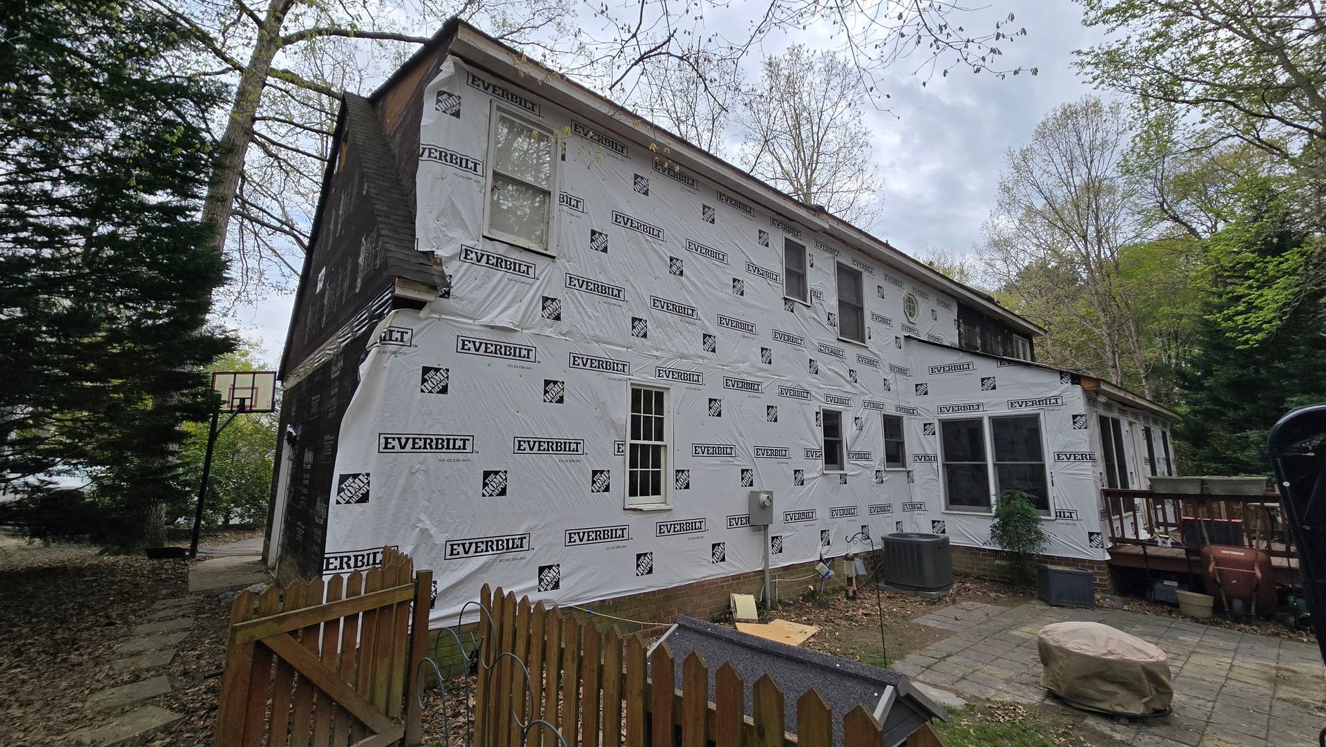 Partially renovated white house with wrapped siding, yard fence, trees, and construction materials