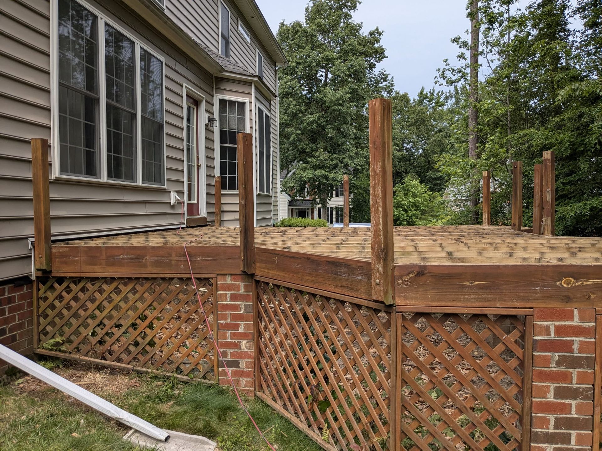 Wooden deck under construction beside a house, with brick lattice skirting and upright support posts.