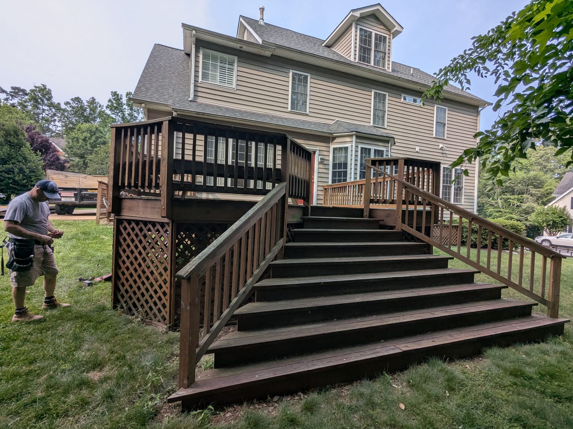 House with large wooden deck and stairs in a grassy yard; a person stands to the left.