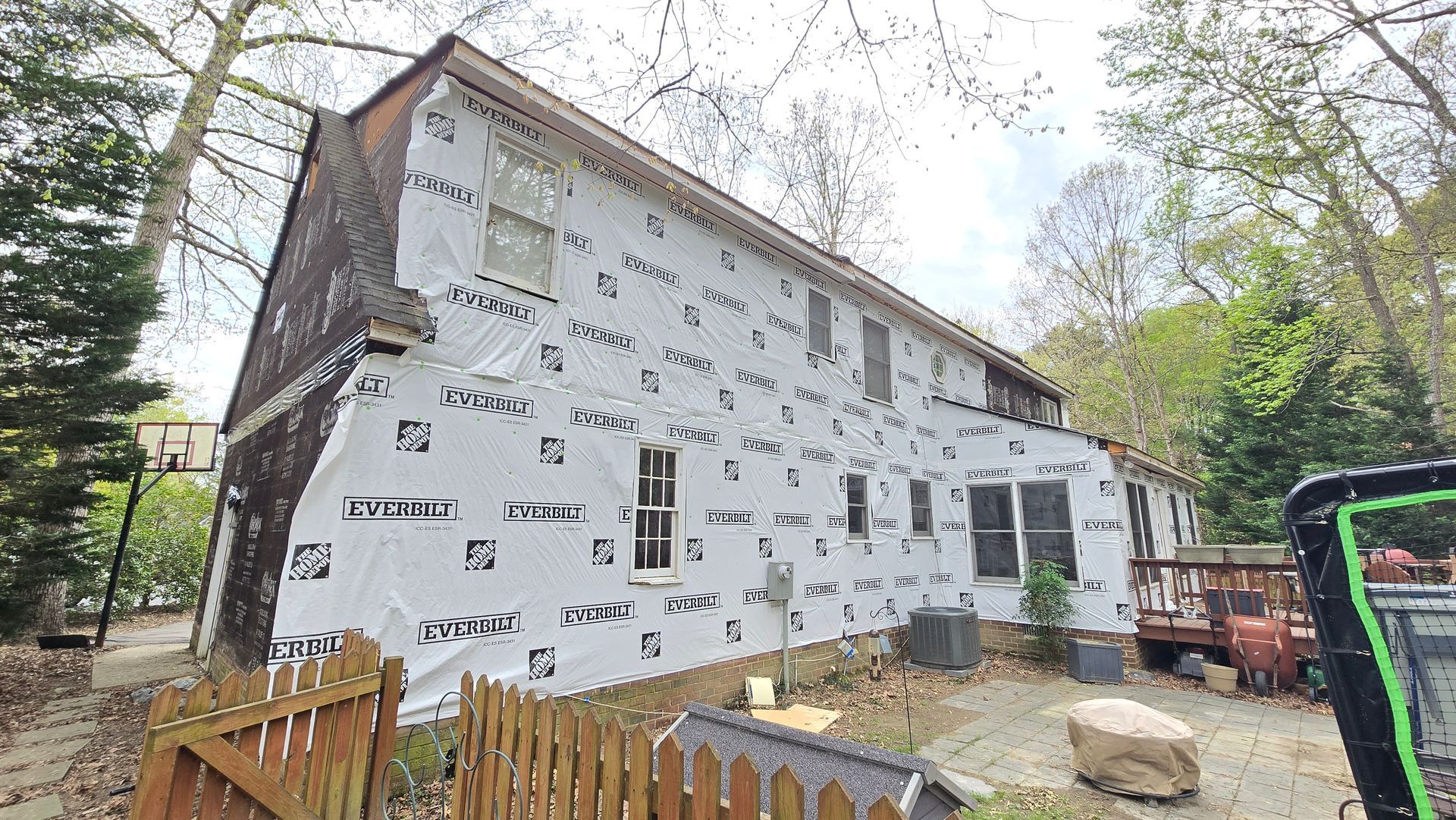 Partially renovated white house with exposed wrap, fenced yard, and trees around it