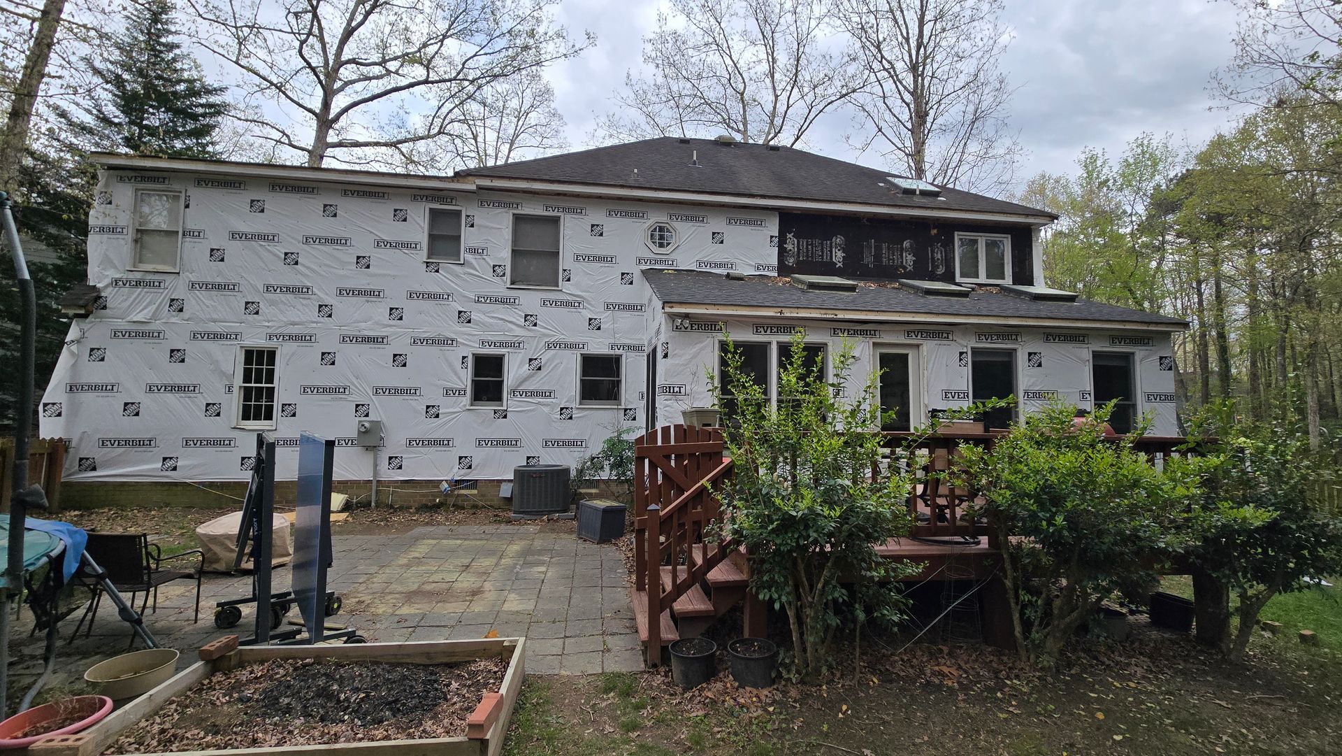 House exterior under renovation, wrapped in white house wrap with exposed windows and a small wooden deck in front.