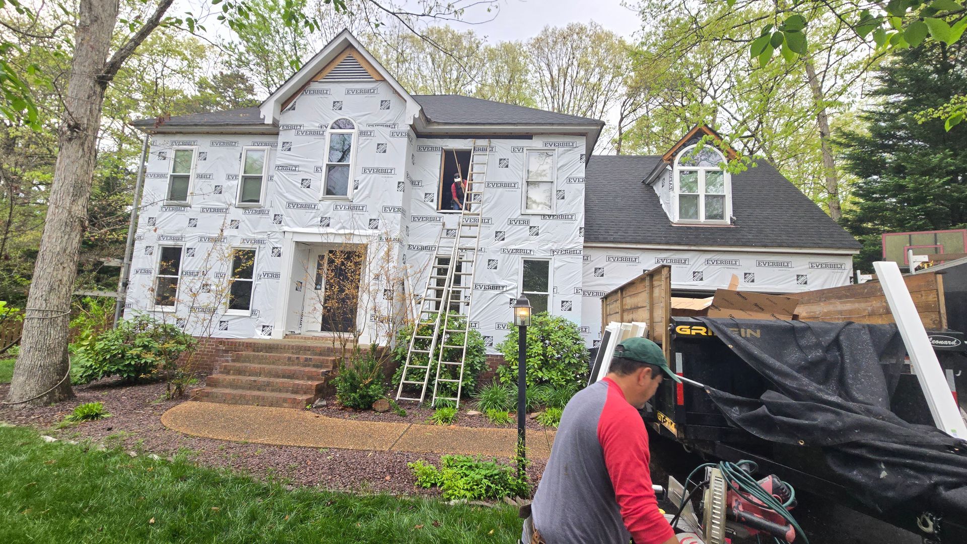 House under renovation with white exterior wrap, ladders, and workers in front yard