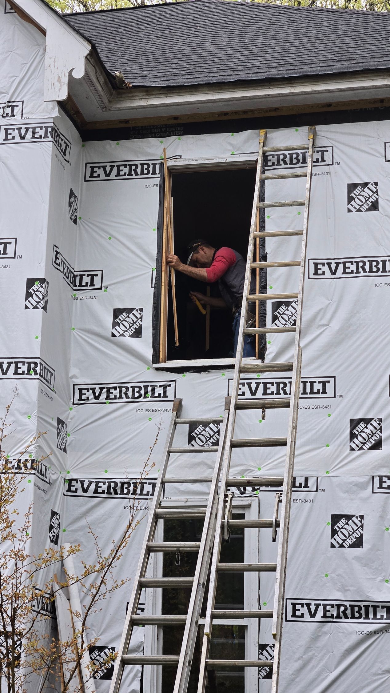 Worker on ladders repairing a house window under renovation, with white wrap covering the exterior.