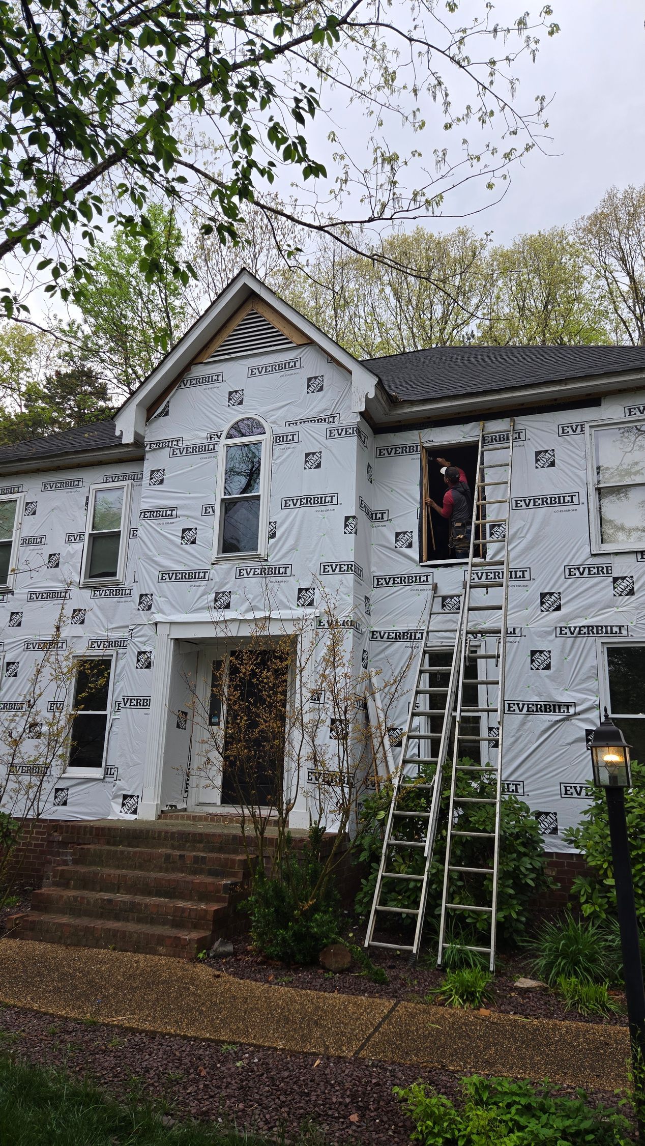 Two-story house under renovation, wrapped in white house wrap with ladders and autumn leaves in front