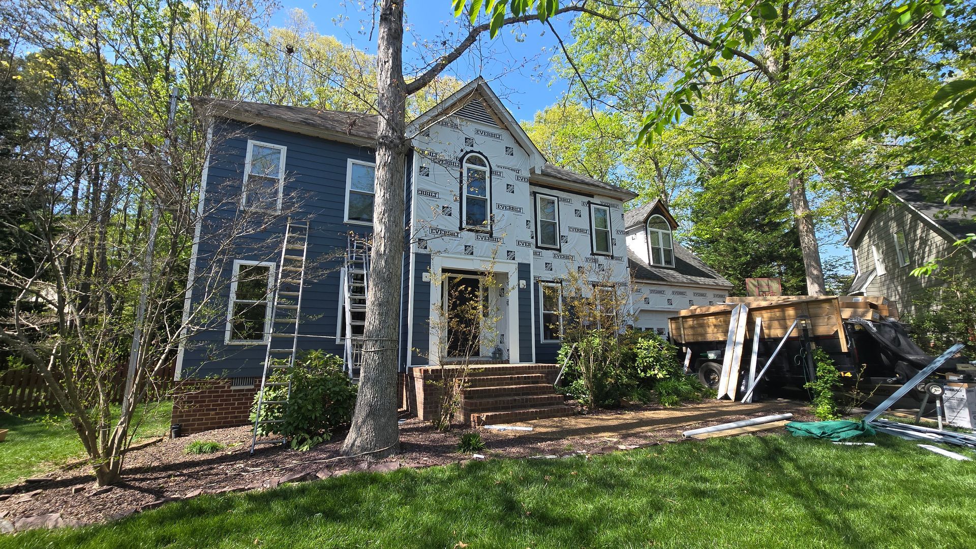 Two-story blue house with white trim, front porch, and a sunlit yard with trees.
