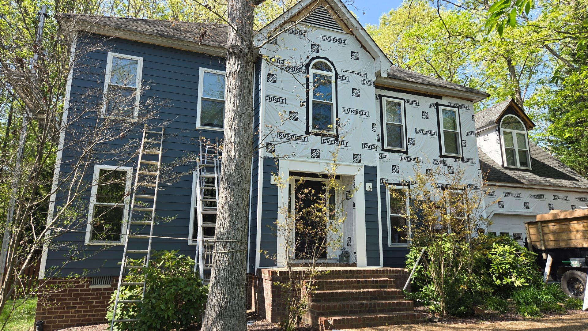 Historic two-story house with dark blue and white siding, front steps, trees, and ladders leaning against it