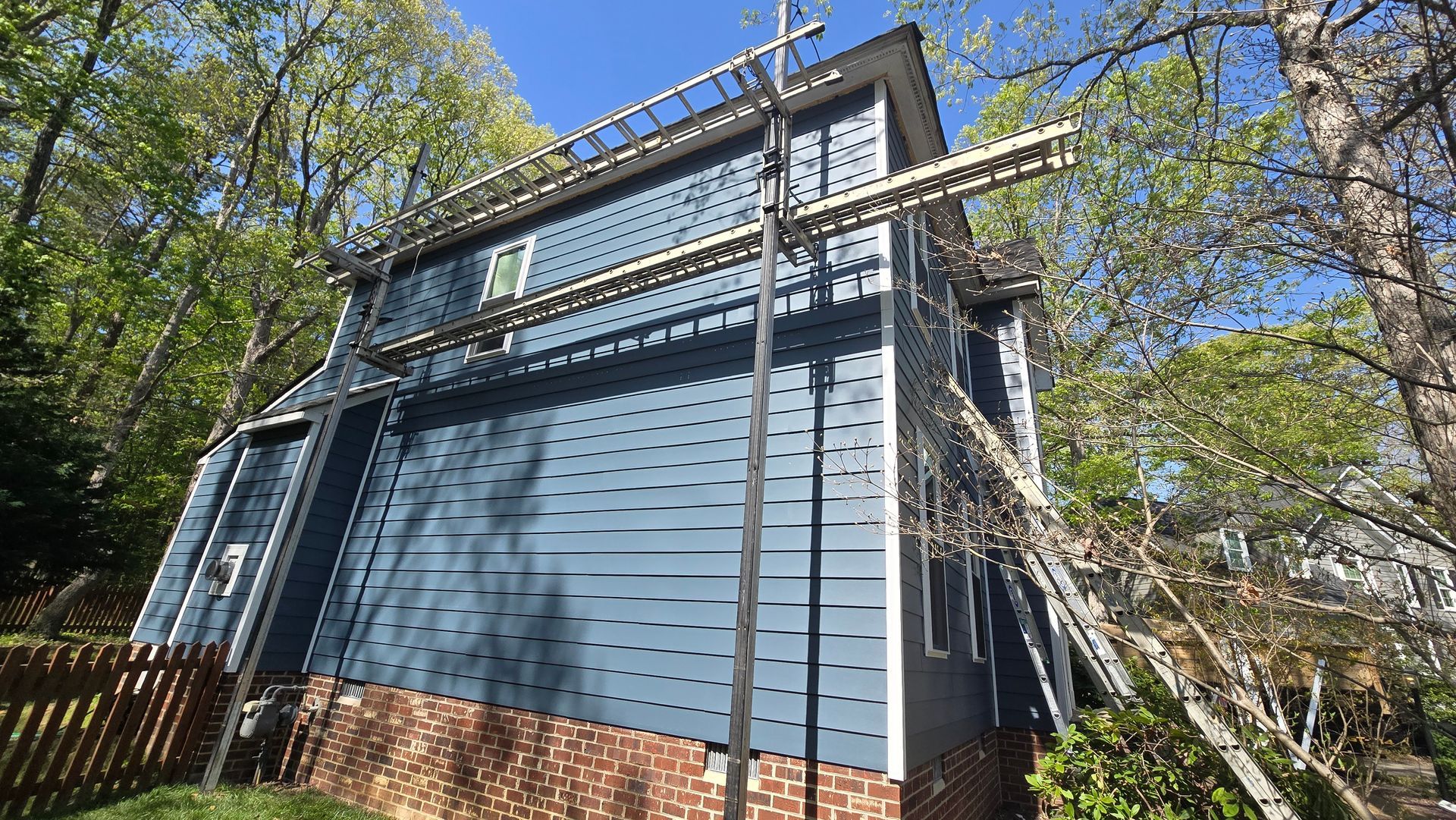 Blue-sided building with external pipes and scaffolding, viewed from below among trees and brick foundation