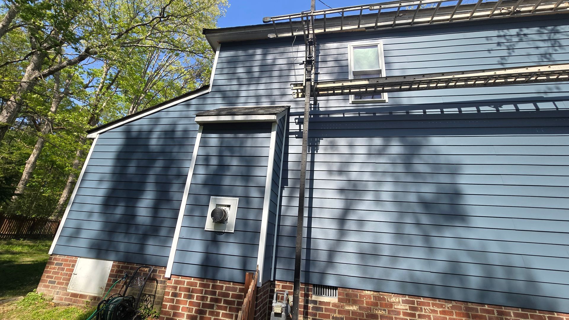 Blue-sided house with brick foundation and utility box, viewed from the side in sunlight.
