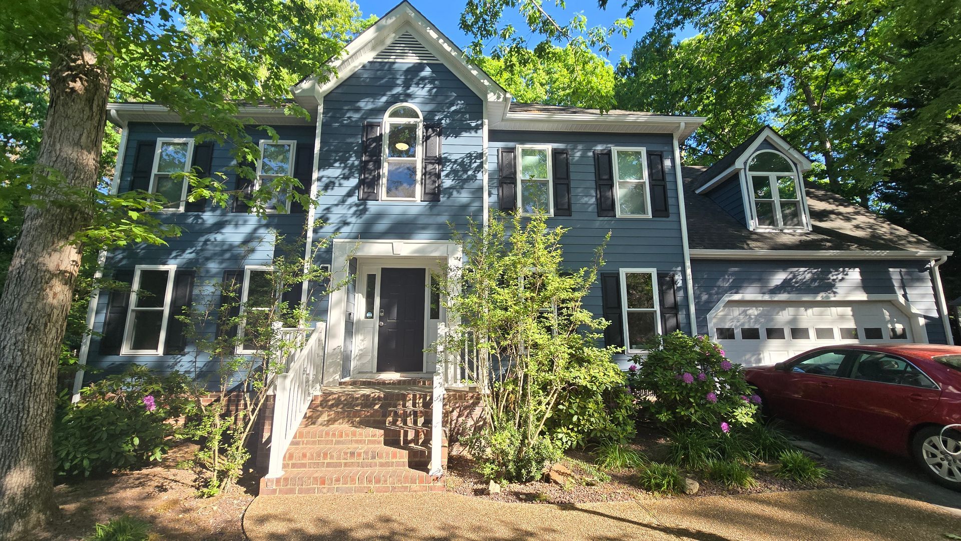 Two-story gray house with white trim, front steps, and a red car in the driveway surrounded by trees.