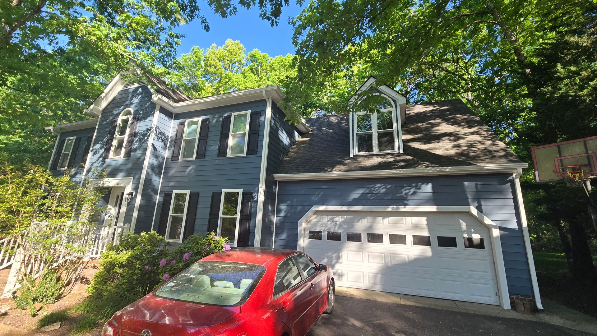 Gray two-story house with white garage, black shutters, and a red car parked in front under trees.