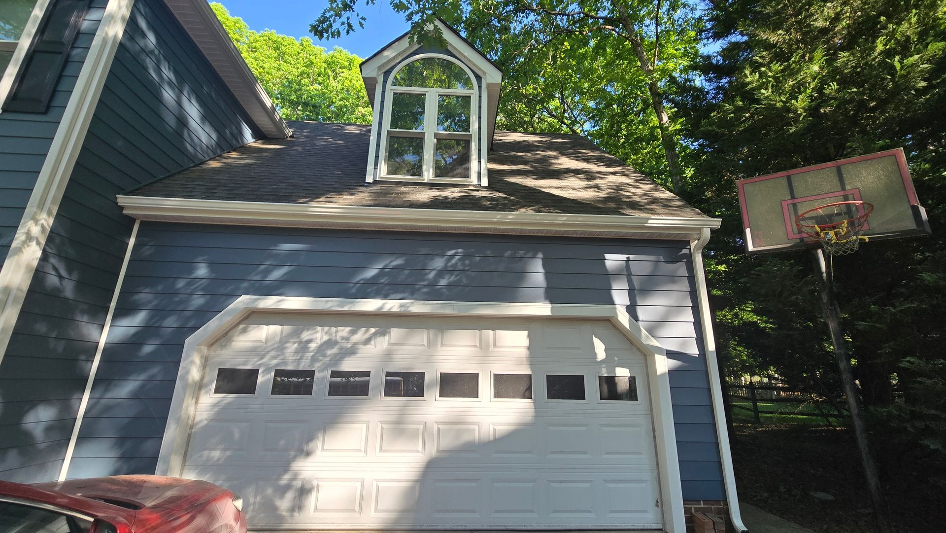 White garage with dormer windows and a basketball hoop beside it, framed by trees and blue sky