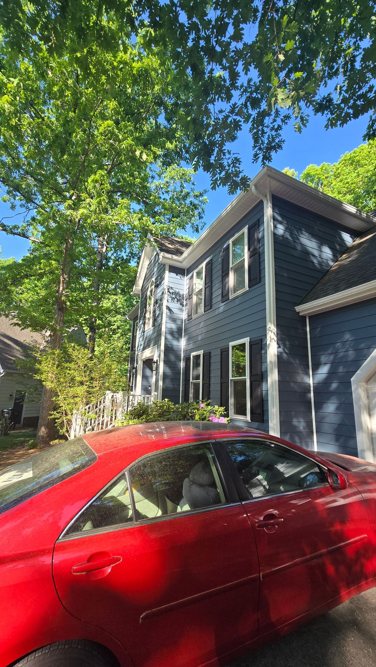 Red car parked beside a gray house under leafy green trees on a sunny day