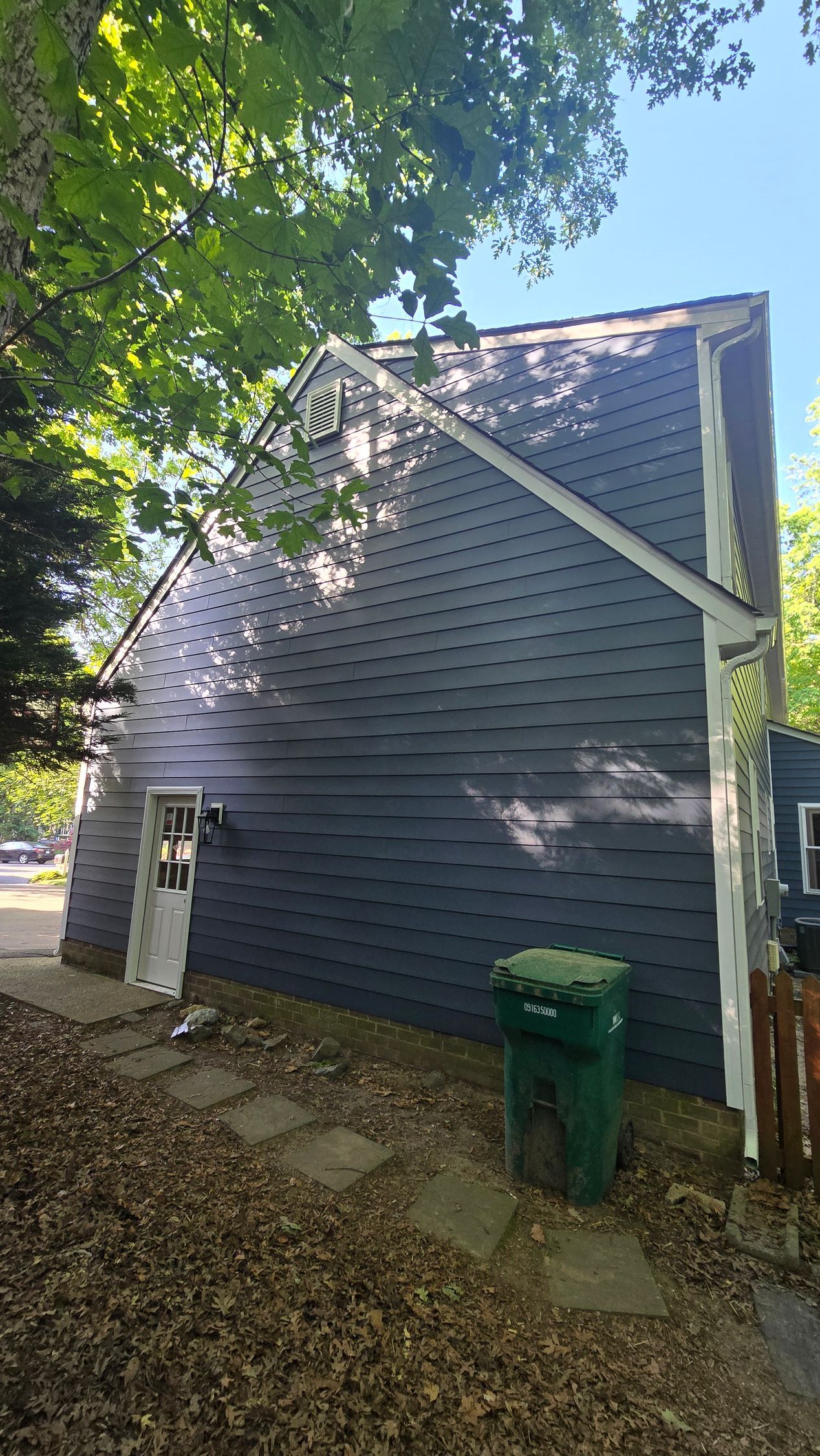 Gray house exterior with diagonal braces, white trim, and green trash bins by a leaf-covered side yard.
