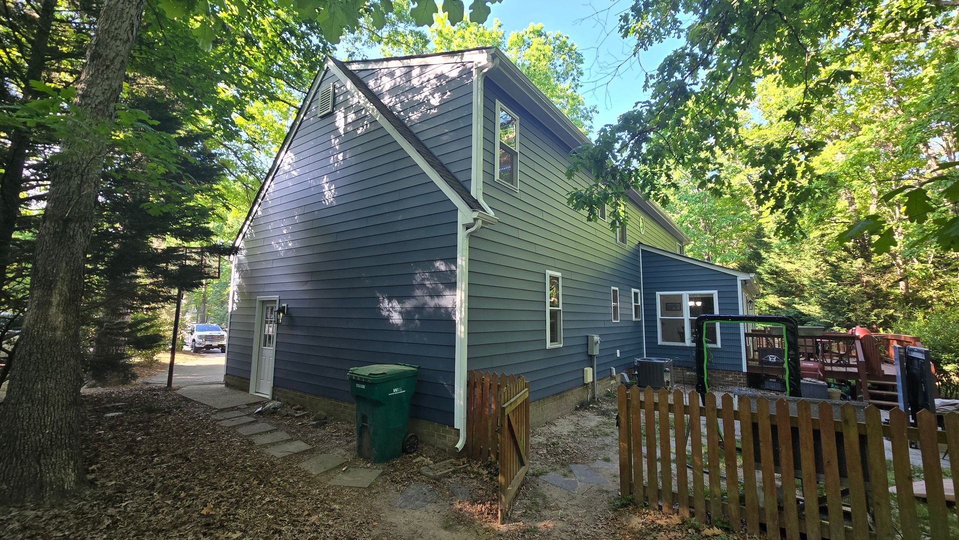 Gray house with a steep roof and trees around it, beside a wooden fence and deck in a wooded yard