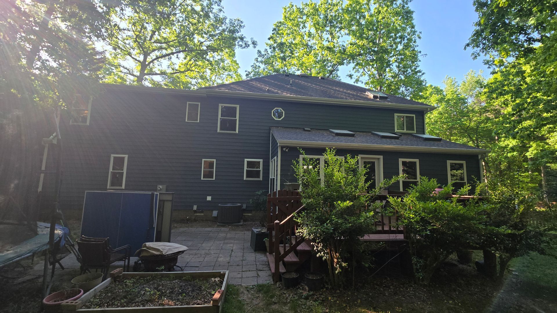 Backyard with a dark two-story house, deck, and lush green trees in bright sunlight