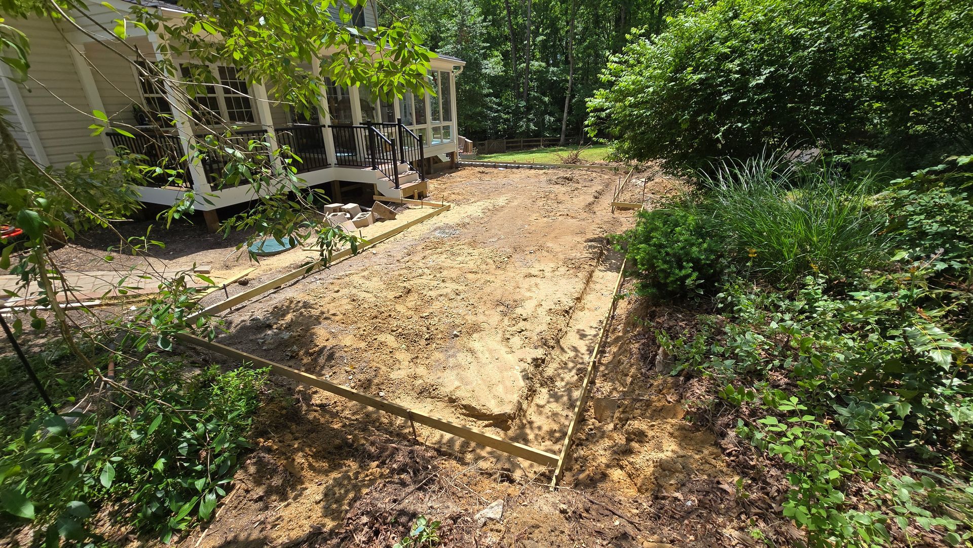 Wooden walkway through a leafy garden beside a porch, with muddy ground and dense green shrubs.