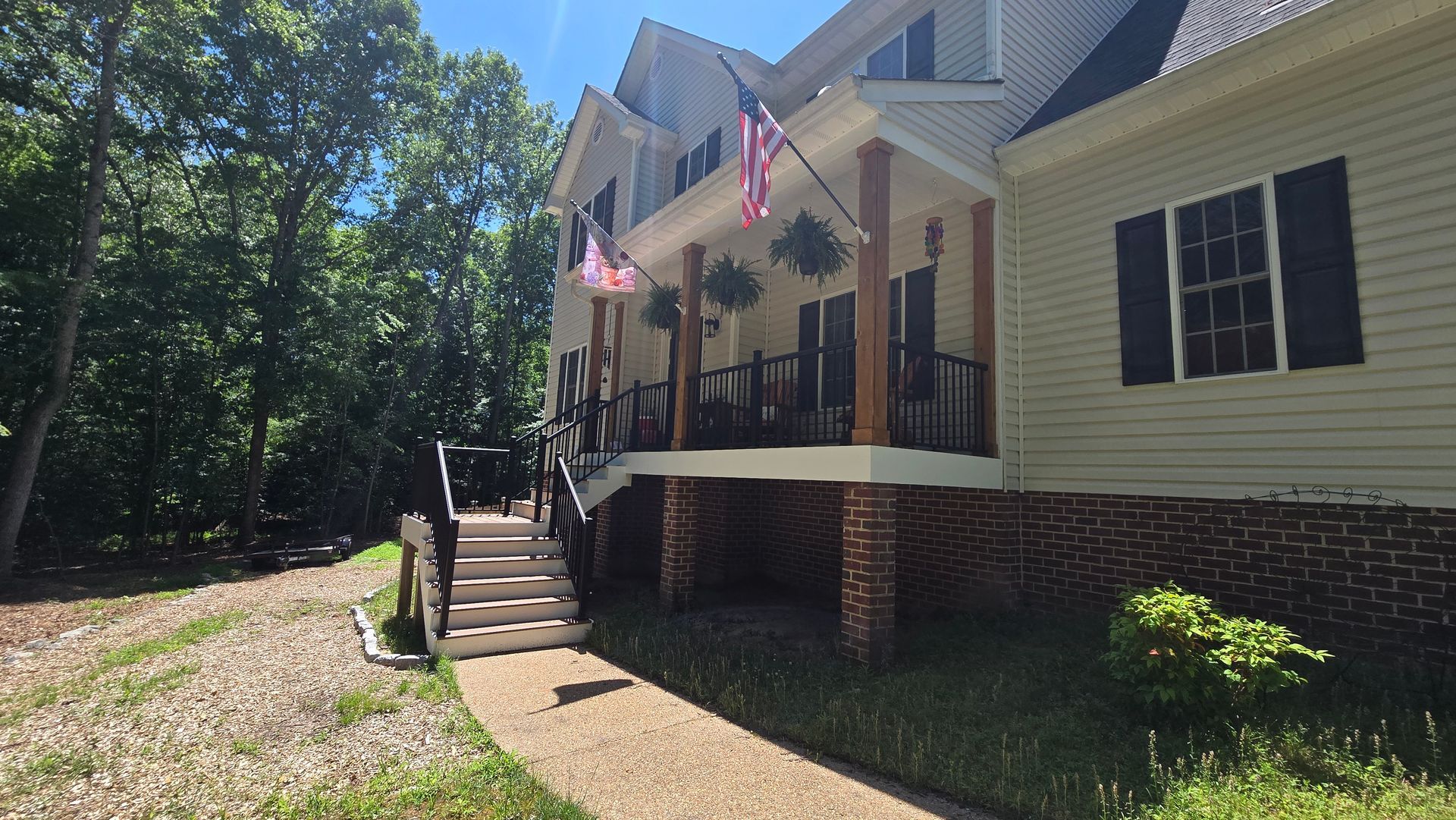 Two-story beige house with front porch, brick foundation, and American flags, viewed from a wooded yard