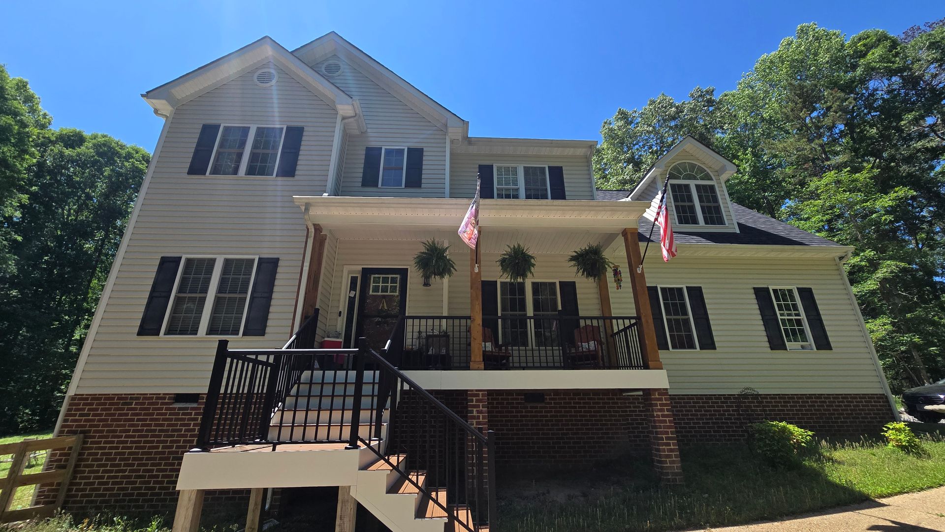 Front view of a light-colored house with black shutters, a porch, and two American flags on a sunny day