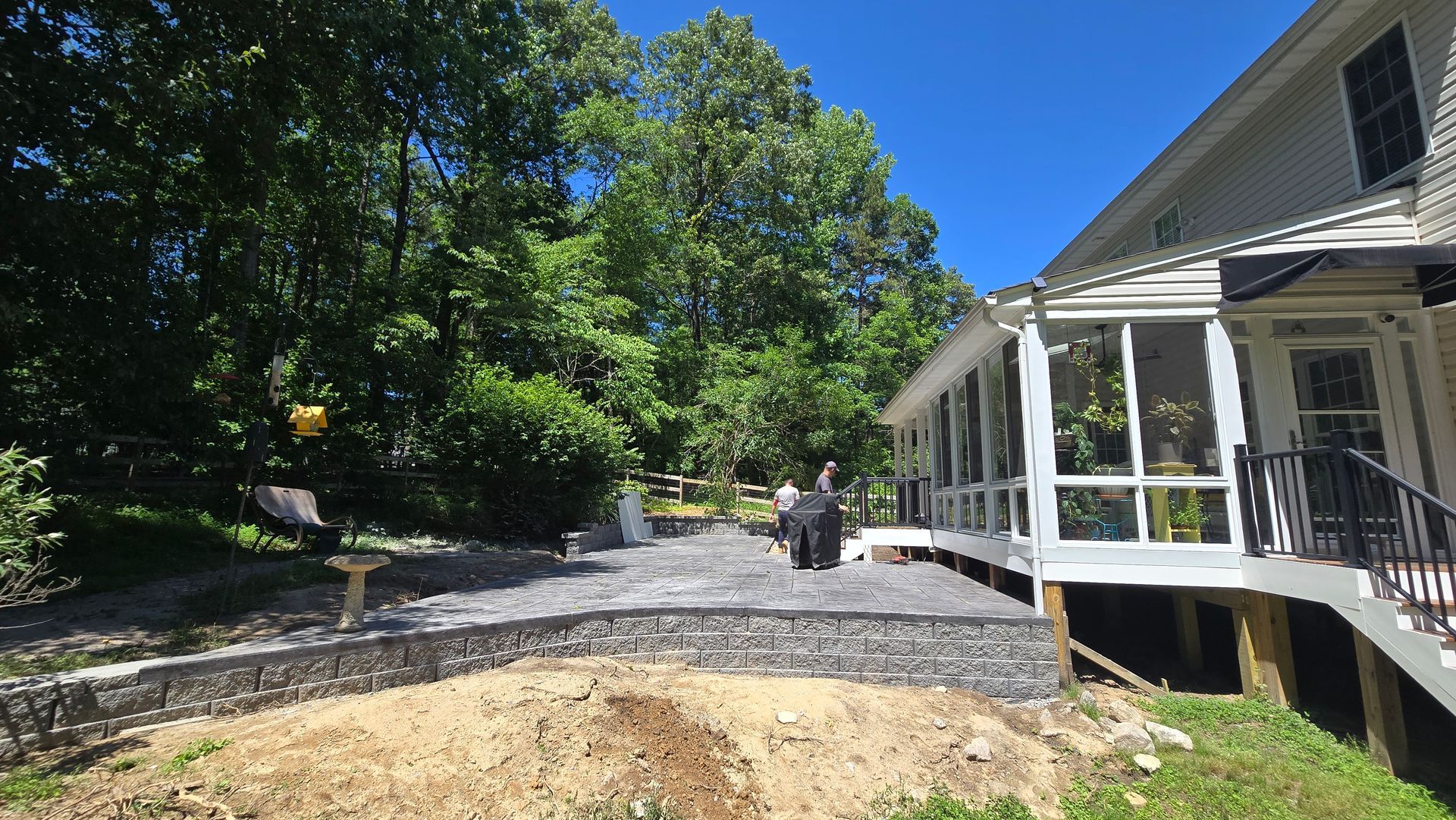Side yard with gravel path, trees, and a house porch under bright sunlight.