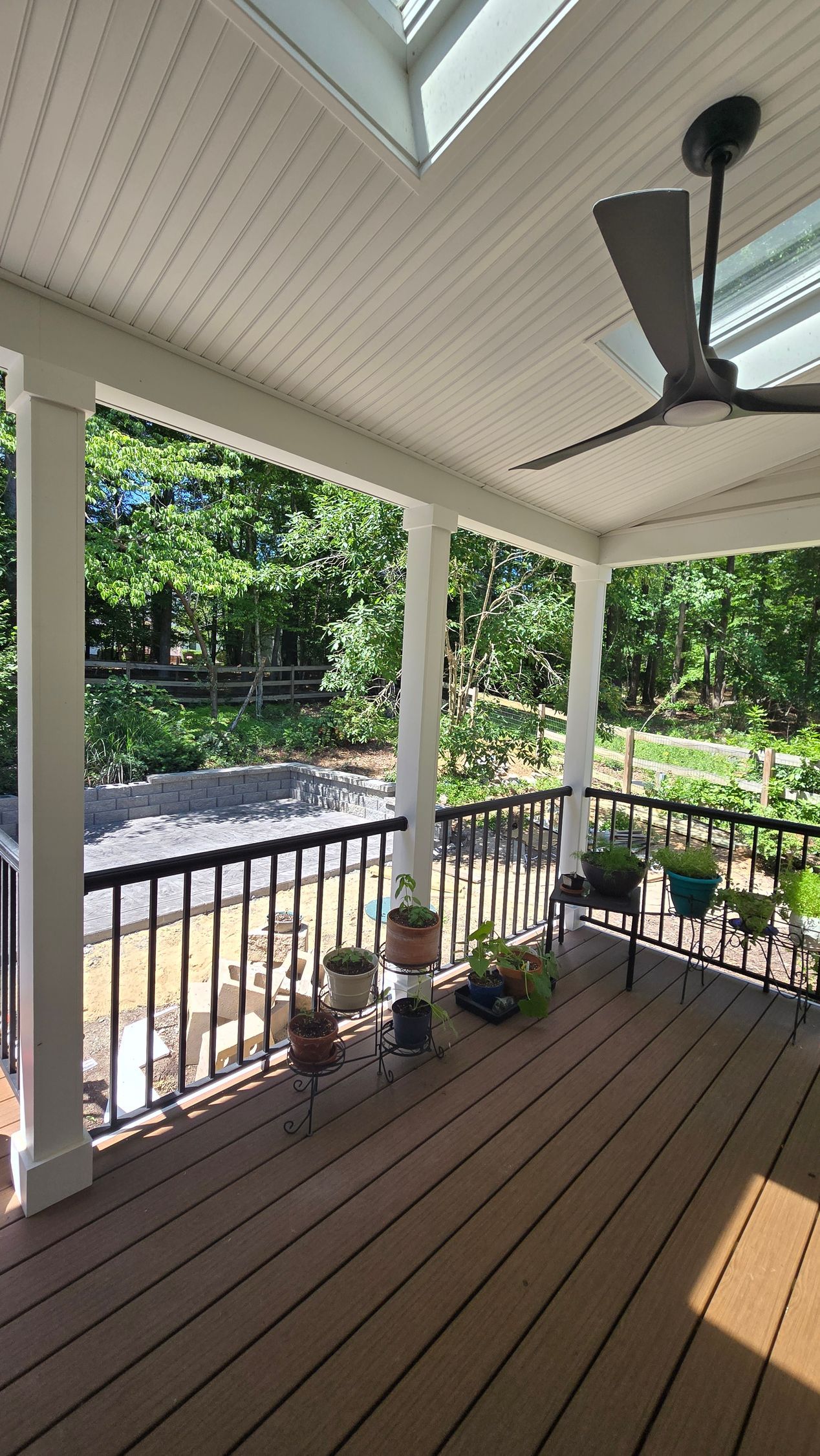 Covered porch with white columns, wooden floor, railing, potted plants, and a ceiling fan overlooking greenery