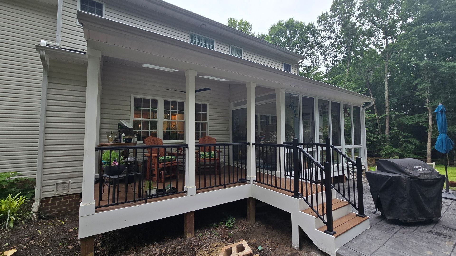 Covered porch of a white house with black railings, potted plants, and a grill on the right side.