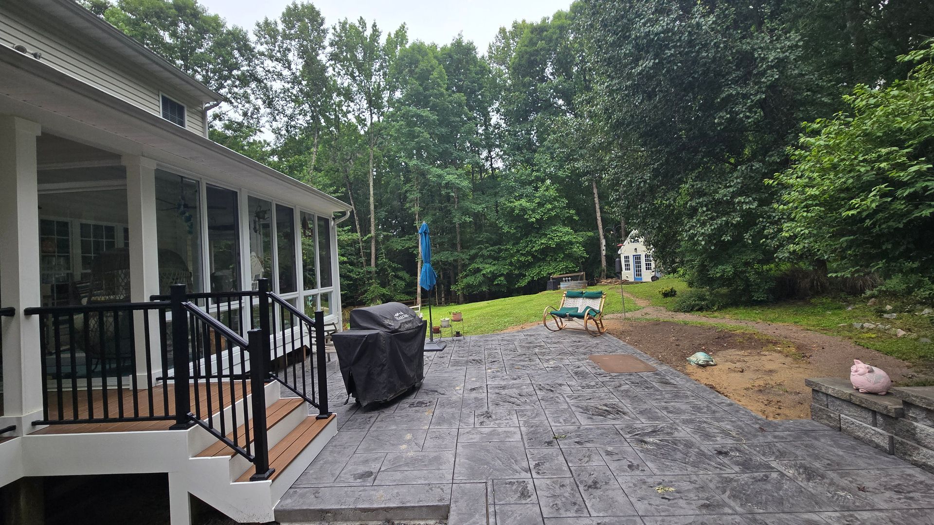 Backyard patio with stone pavers, grill, and chairs beside a house and wooded yard