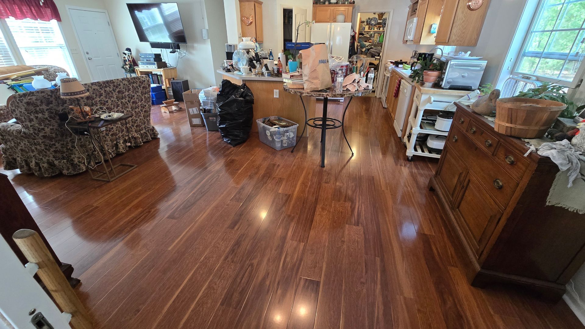 Cluttered living room with polished wood floors, furniture, boxes, and a bright kitchen in the background