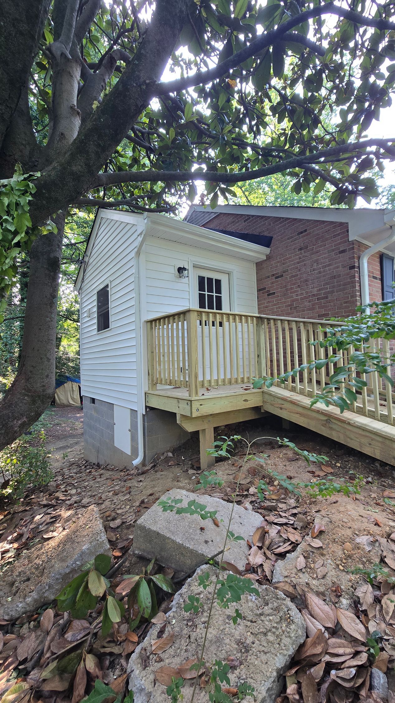 Small white playhouse with a wooden ramp and deck among trees and rocks in a backyard