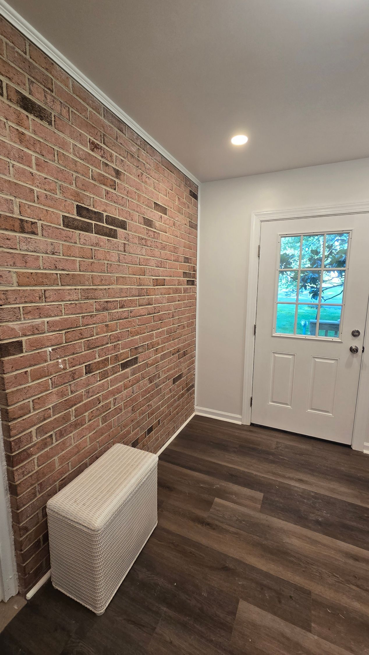 Small entryway with exposed brick wall, white front door, dark wood floor, and a wicker storage basket.
