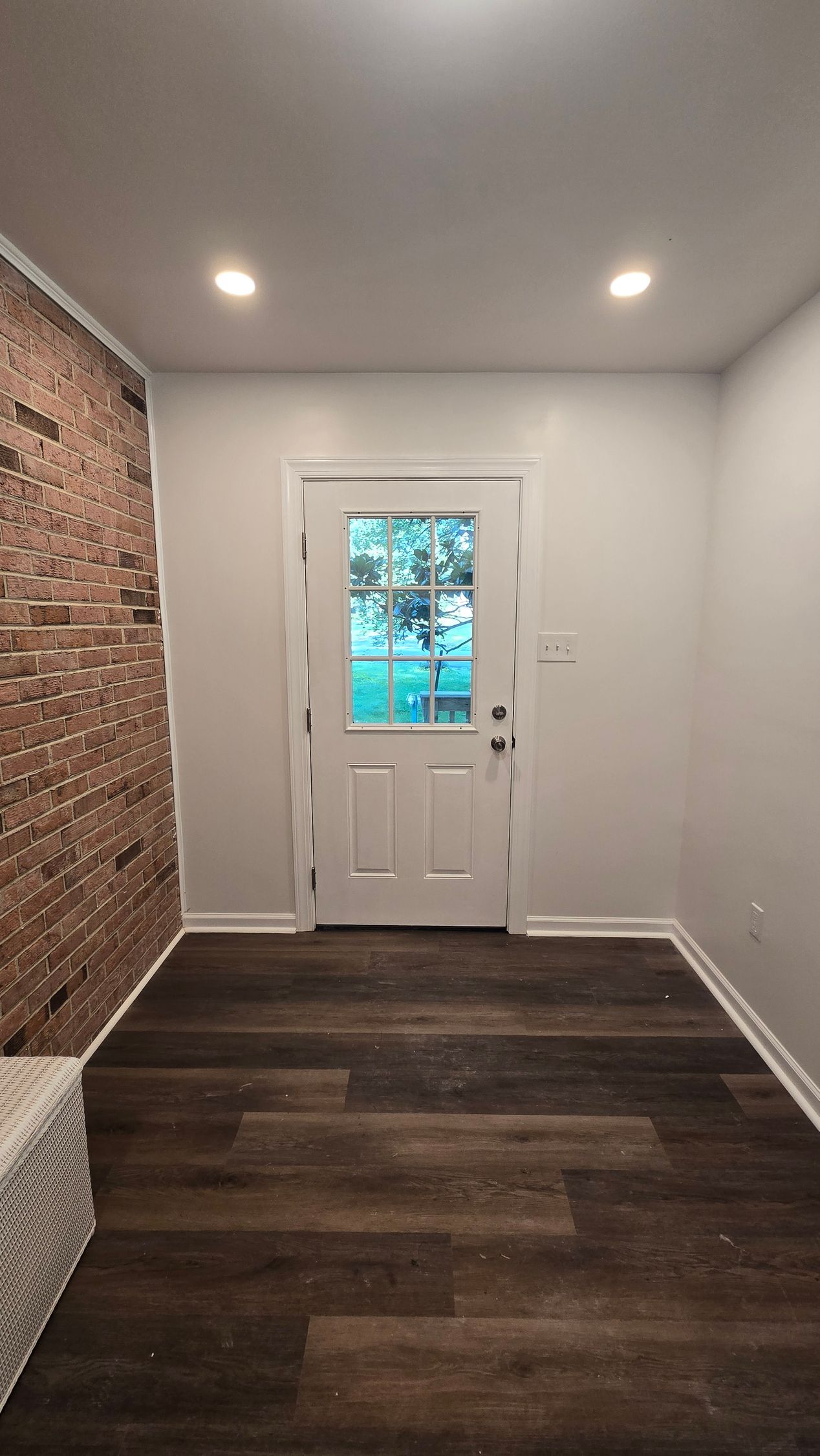 Empty narrow room with dark wood floor, white walls, exposed brick accent, and a door with blue light outside.