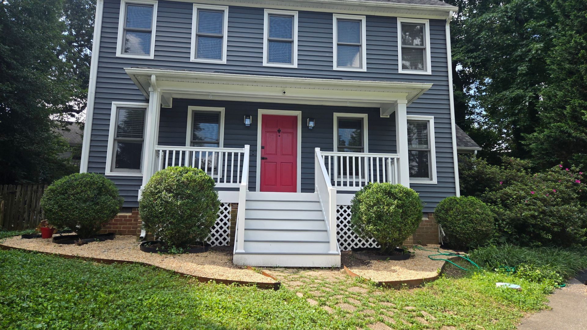 Two-story gray house with white trim, red front door, and small front porch with steps and shrubs