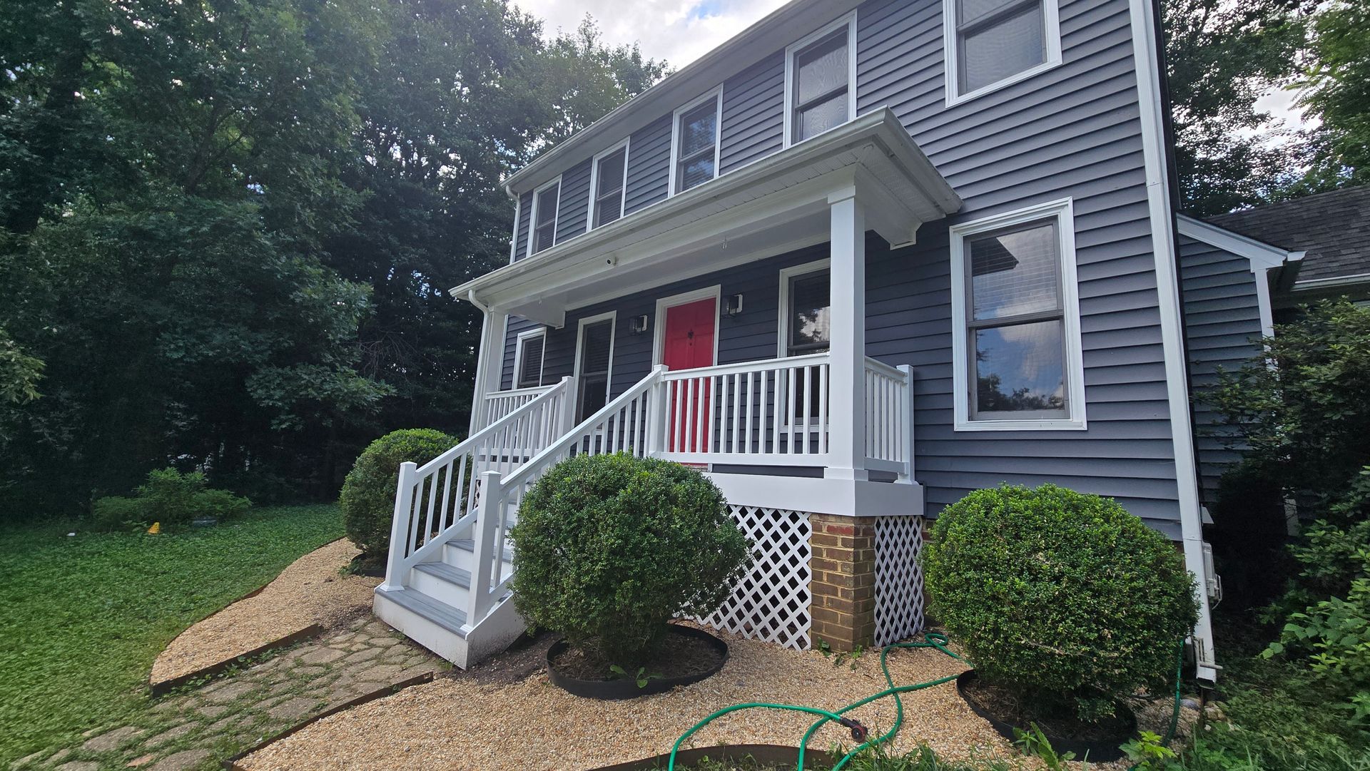 Gray two-story house with white porch, red front door, and stone skirting beside a green lawn.