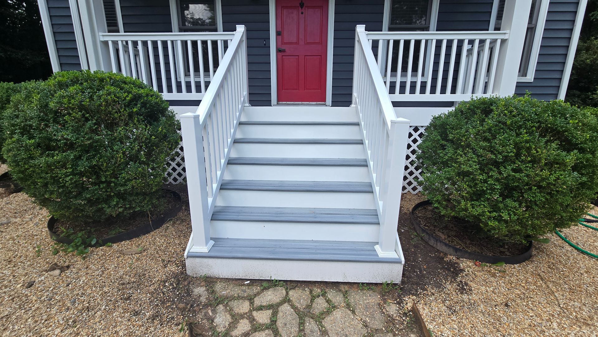 Front porch with white steps and railings leading to a red front door, flanked by two green bushes.